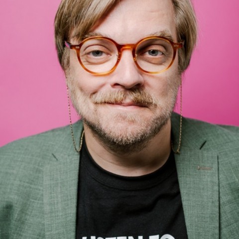 Nilz Bokelberg with glasses, blond hair and beard smiling in front of pink wall