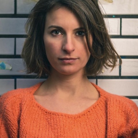 Nora steht vor einer hellen Wand und trägt einen orangefarbenen Pullover