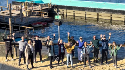 Das re:publica-Team steht auf dem Sandstrand am Ufer vor dem Badeschiff in der Sonne und winkt.