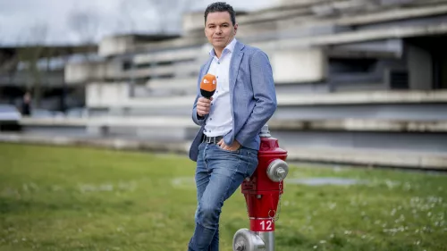 Özden steht auf einer Wiese und lehnt sich an einen Hydranten. Er hält ein Mikrofon in der Hand.