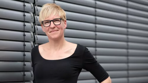 Dies ist ein Bild von Eva Wolfangel. Sie trägt eine Brille, kurze Haare und steht vor einer Jalousie.