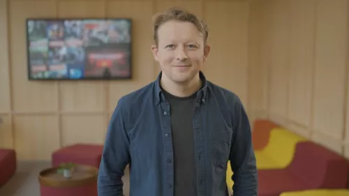 Philipp Lorenz-Spreen trägt kurze Haare, ein Hemd und darunter ein T-shirt. Im Hintergrund ist verschwommen eine Sofagarnitur und ein Bildschirm zu sehen. Er lächelt in die Kamera