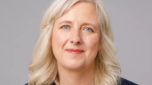 Foto von Carole vor einem grauen Hintergrund. Sie trägt offene, gestufte Haare und lächelt.