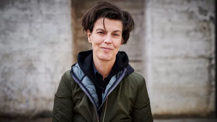 Carolin Emcke trägt eine grüne Jacke und kurze Haare. Sie steht vor einer Wand.
