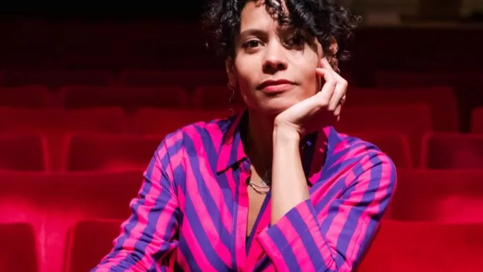 Auf dem Foto ist Emilia Roig in einem Theater- oder Kinosaal zwischen roten Vorstellungssesseln zu sehen. Sie trägt ein gestreiftes Oberteil und blickt direkt in die Kamera.