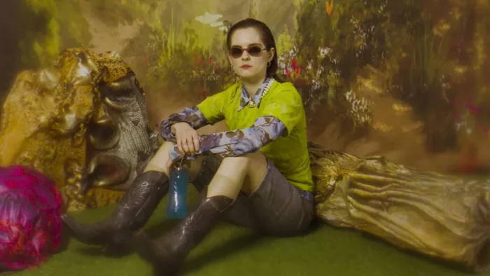 Die Kunstfigur Fuffifufzich sitzt in Cowboystiefeln auf dem Boden. Sie trägt eine Sonnenbrille. Die Leinwand hinter ihr sieht aus wie ein Gemälde. 