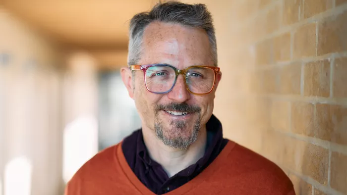 Sam Gregory trägt eine Brille, kurze Haare und Bart. Er trägt einen Pullover und darunter ein Hemd. Er lächelt in die Kamera und lehnt an einer Backsteinwand.
