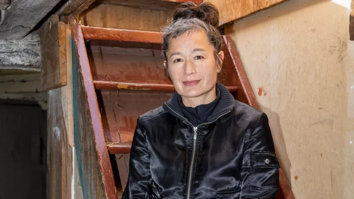 Hito Steyerl sitzt auf einer Treppe in einem Keller. Hito trägt einen Zip-Hoodie und Dutt. Sie schaut in die Kamera.