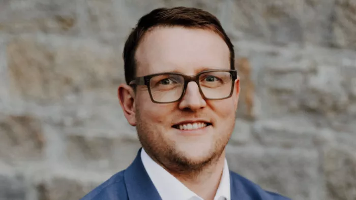 Foto von Matthias Quent. Er steht vor eine Steinwand und lächelt in die Kamera. Dazu trägt er kurze Haare, Anzug und Brille.