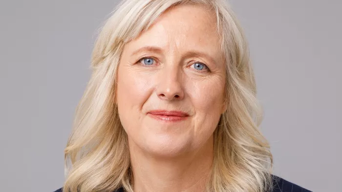 Foto von Carole vor einem grauen Hintergrund. Sie trägt offene, gestufte Haare und lächelt.