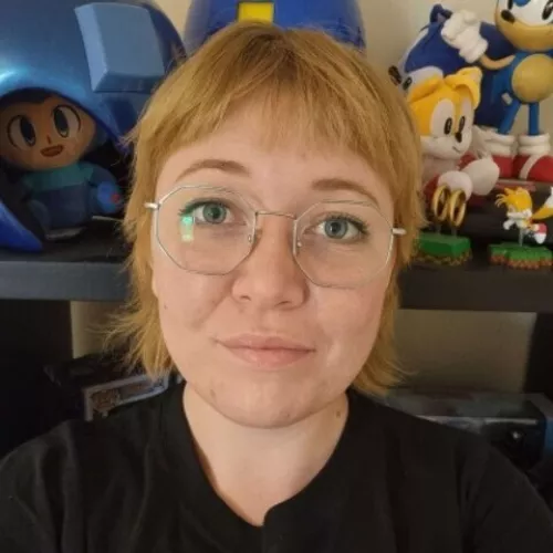 Headshot of Sky, a person with short blond hair and glasses standing in front of a shelf with colorful toys.