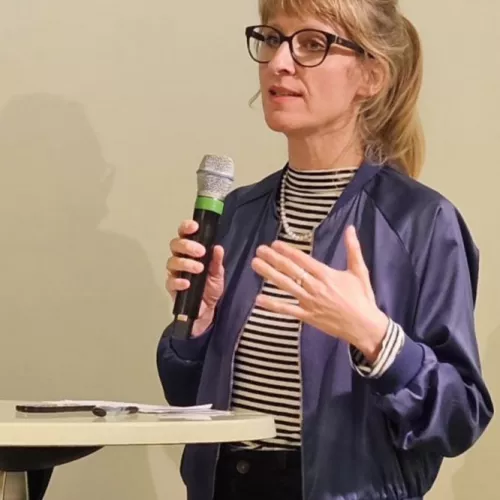 Eine blonde Frau mit dunkler Brille steht an einem Pult und hält ein Mikrofon in der Hand und spricht gerade