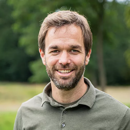 Portraitfoto von Christoph Schmitz lachend im Grünen mit khakifarbenen T-Shirt