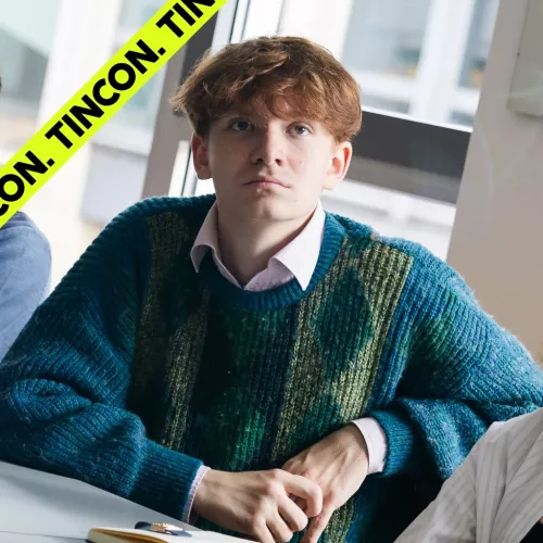 Junger Mann mit roten Haaren, rosafarbenen Hemd und blauem Strickpullover, schaut konzentriert über die Kamera hinweg