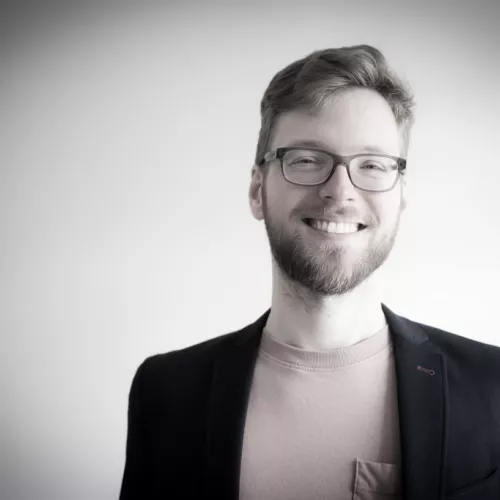 Mann mit Brille, Bart und breitem Lächeln in einem dunklen Jacket und rosa Shirt vor einem hellen Hintergrund