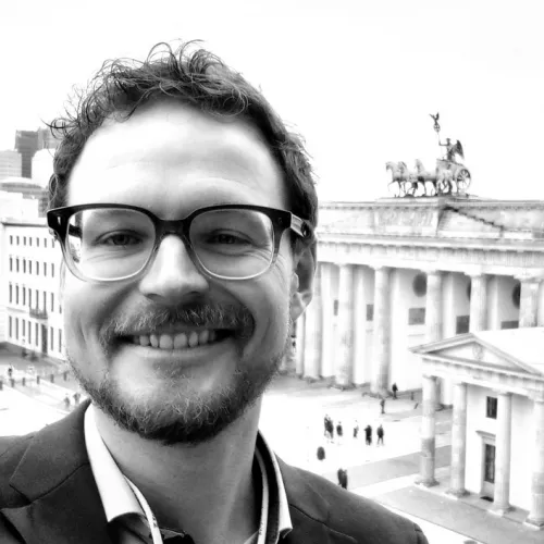 Ein lächelnder Mann in dunklem Anzug und Hemd in halbnaher Aufnahme ist vor dem Pariser Platz und dem Brandenburger Tor zu sehen.