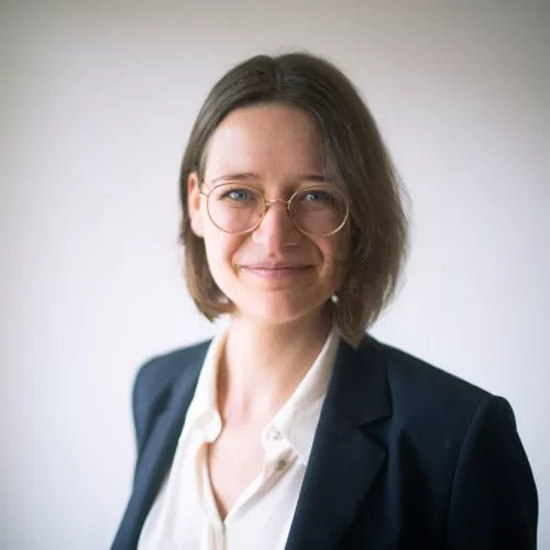 Auf dem Bild ist Julia Bringmann zu sehen. Sie trägt ein weißes Hemd, ein dunkles Jacket und eine Brille.