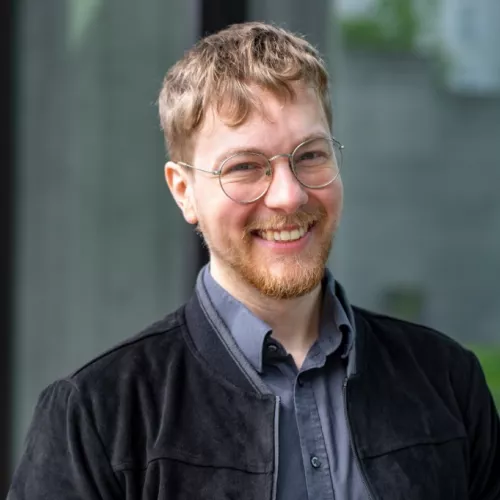 Ein Bild von Thomas Laschyk,  lächelnd vor einer Glaswand. Er trägt eine runde Brille, ein dunkelgraues Hemd und eine schwarze, samtartige Jacke. Im Hintergrund ist ein unscharfer, moderner Außenbereich mit Glas und etwas Grün zu erkennen
