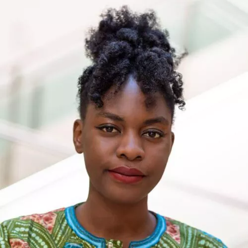 Esther Mwema, black African woman with curly afro hair in a bun, wearing green and red African material shirt, stands against blurred light background.