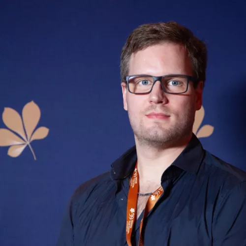 Portrait von Christian Zipfel. Junger Mann mit Brille, der vor der Fotoleinwand des Warsaw International Film Festival steht. 