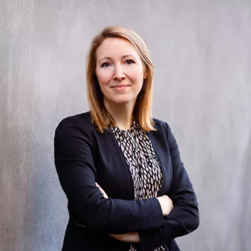 Eine erwachsene Person steht vor einer hellgrauen Betonwand. Sie trägt einen dunklen Blazer über einer gemusterten Bluse und hat die Arme vor dem Körper verschränkt. Das schulterlange, helle Haar ist offen getragen. Die Person blickt direkt in die Kamera und lächelt leicht. Der Hintergrund ist schlicht und unscharf, wodurch die Person im Vordergrund steht.