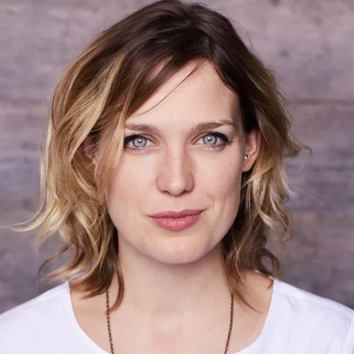 Cori ist Mitte 30 und Irin. Sie hat hellblaue Augen und dunkelblondes Haar. sSie trägt ein weißes T-shirt.
