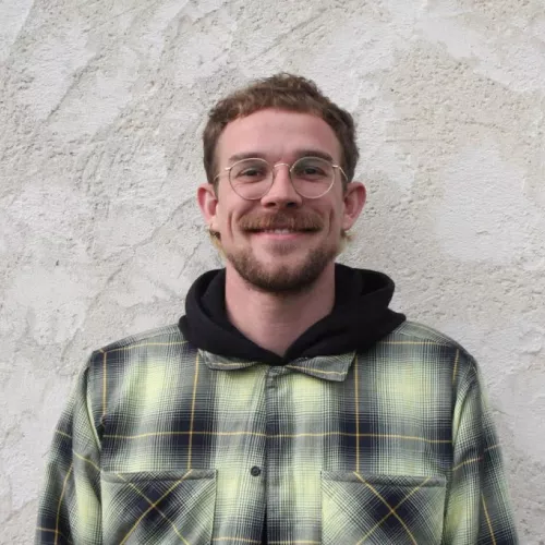 Max steht vor rau verputzter Wand, trägt Hoodie und gelb schwarz karierte Jacke. Klassischer Portrait Fotoaufbau. Er grinst.