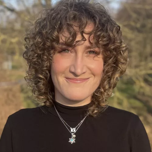 Frau mit blonden Locken lächelt in die Kamera. Sie trägt ein schwarzes schlichtes Oberteil und eine Kette mit Davidstern.