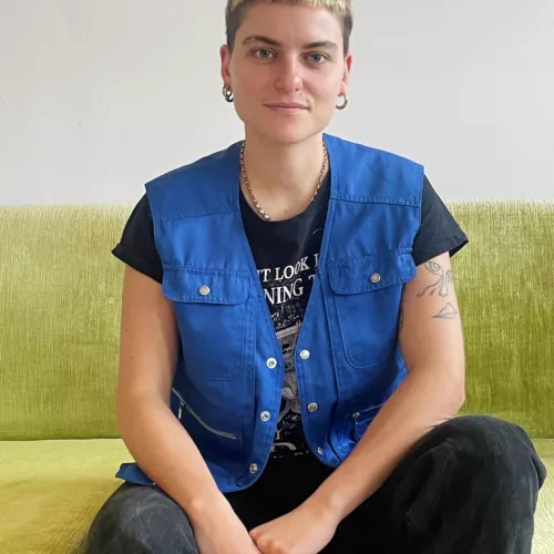 Taiina sitzt auf einem hellen Samtsofa und trägt eine schwarze Jeans und eine blaue Weste.