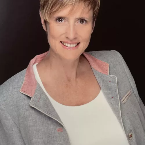 Porträtfoto einer lächelnden Frau mit kurzem blondem Haar vor dunklem Hintergrund. Sie trägt eine helle Jacke und ein weißes Oberteil.