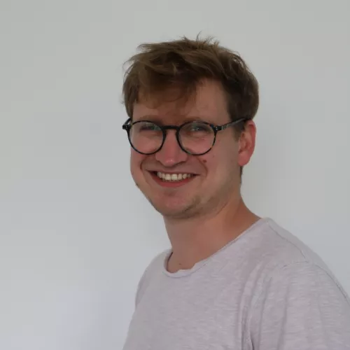 David Hartmann mit einer runden Brille und einem hellen T-Shirt lächelt in die Kamera. Der Hintergrund ist schlicht weiß, und das Bild zeigt eine freundliche, entspannte Ausstrahlung.