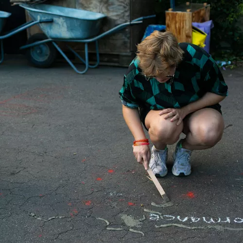 Iz, a white nonbinary person with short blonde hair who is wearing a green shirt and sunglasses, is kneeling on the ground during a performance of their arts-design duo MELT. They are using a stick to pour concrete into a crack on the floor.