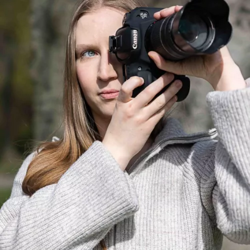 Jana steht in der Natur vor einem Baum. Sie trägt einen beige farbenden Pullover und hat ein Kamera vor ihrer rechten Gesichtshälfte. Ihre langen Haare fallen über die linke Schulter.