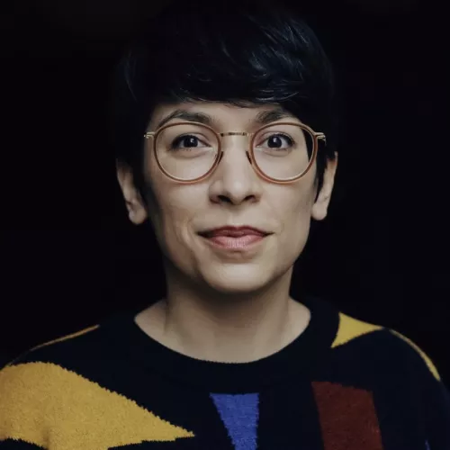 Eine Frau mit runder Brille und kurzen schwarzen Haaren schaut in die Kamera.