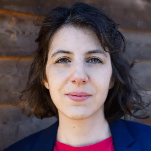 Das Bild zeigt ein Porträt der lächelnden Juliane Vicari mit dunklen Locken, magentafarbenem Shirt und blauem Blazer vor einer Holzwand.