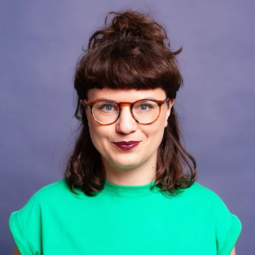 Das Bild zeigt einen Headshot von Lilith Boettcher, mit Brille, braunen Haaren und einem grünen Tshirt.