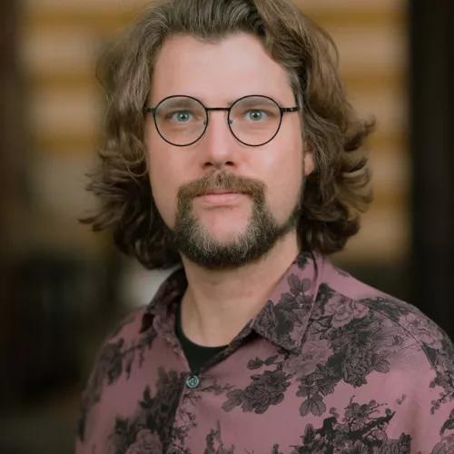 cis-Mann, bärtig, langes Haar, runde Brille, gemustertes Hemd