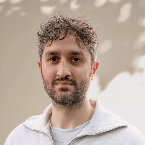 Ein Mann Anfang dreißig mit dunkel braunen Haaren, einem Drei-Tage-Bart und braunen Augen. Er trägt ein hellgraues Sweatshirt und steht vor einer gelblichen Wand. Das Licht der Sonne fällt durch die Blätter eines Baumes auf die Wand.