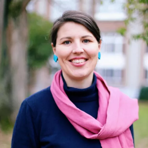 Demale smiling to the camera. Blue sweater with pink scarf and blue earrings. building in the background.