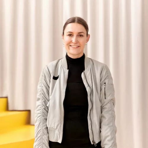 Das Bild zeigt ein Portrait von Franziska Lindner. Sie ist bis zur Hüfte zu sehen. Sie hat braune, streng zu einem Zopf gebundene Haare, ist circa 30 Jahre alt und lacht in die Kamera. Ihre Arme hängen locker nach unten. Sie trägt ein schwarzes Oberteil und eine graue Bomberjacke. 
