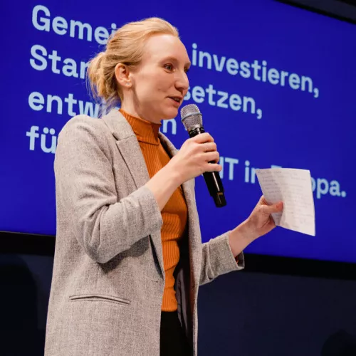 Speakerin in grauem Blazer, orangem Rollkragenshirt. Blonde Haare, zu einem Zopf gebunden. Ein Mikro in der rechten Hand, Moderationskarten in der linken.