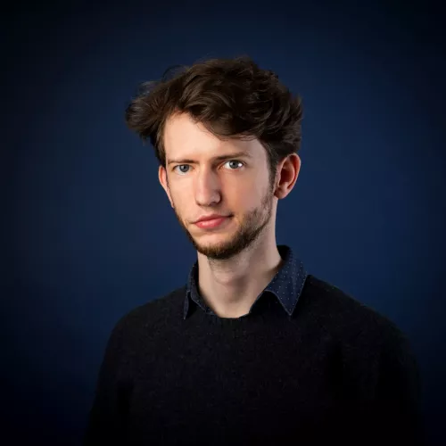 Ein junger Mann Ende 20 vor einem dunkelblauen Fotohintergrund. Er hat braune Haare und leichen Bartwuchs, sowie einen schwarzen Pullover an. Er lächelt leicht in die Kamera. 