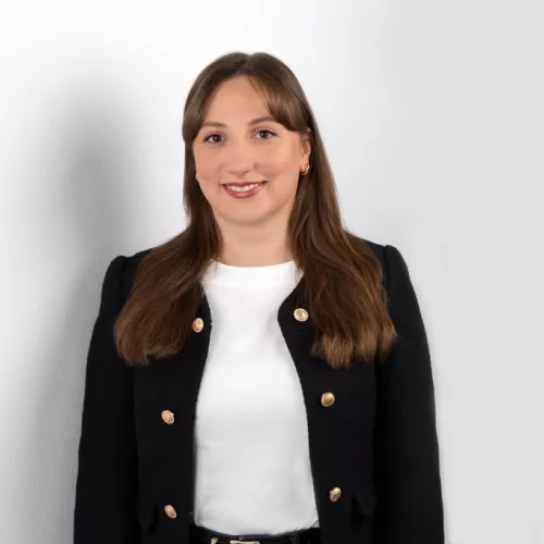 Eine junge Frau steht vor einer hellen Wand und lächelt in die Kamera. Sie trägt ein weißes Oberteil und eine schwarze Jacke mit goldenen Knöpfen.