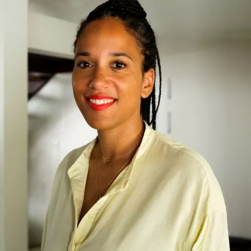 A woman with braided hair pulled back stands indoors, facing the camera with a slight smile. She is wearing a light-colored blouse and a delicate necklace. The background is minimal, with a white wall and a wooden staircase partially visible, creating a calm, modern interior setting.