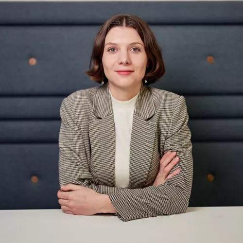 Porträt einer weiblichen Person, die mit verschränkten Armen und einem Blazer auf einem Sofa sitzt