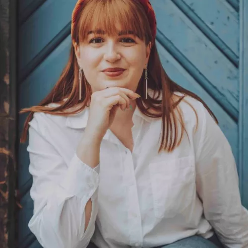 Das Bild zeigt eine junge Frau, die vor einer blauen Holztür sitzt. Sie hat langes, glattes, rötliches Haar mit Pony und trägt ein auffälliges, orangefarbenes Haarband. Sie hat helle Haut und ein warmes Lächeln. Die Frau stützt ihren Kopf leicht auf eine Hand, während sie in die Kamera schaut. Sie trägt eine weiße Bluse mit aufgerollten Ärmeln und eine hellblaue Jeans. Ihre Ohrringe sind lang und dekorativ. Der Hintergrund besteht aus einer diagonal gemusterten Holztür in k