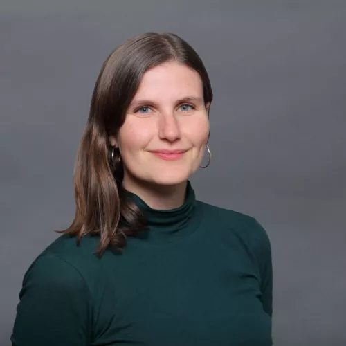 Man sieht eine weiblich gelesene Person im Portraitformat vor grauem Hintergrund. Sie trägt einen petrolfarbenes Langarmshirt und goldene Kreolen. Sie lächelt.   