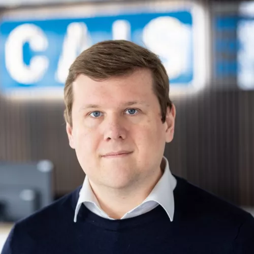 Ein weißer mitteleuropäischer Mann mit kurzem Haar, der in die Kamera guckt und einen dunkelbauen Pullover trägt. Im Hintergrund sieht man das Logo von CAIS.