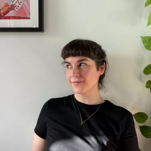 dark-haired woman sitting against a white wall, framed by a picture to her left and a plant to her right. she's looking off to the side with a soft smile and wearing a black shirt.