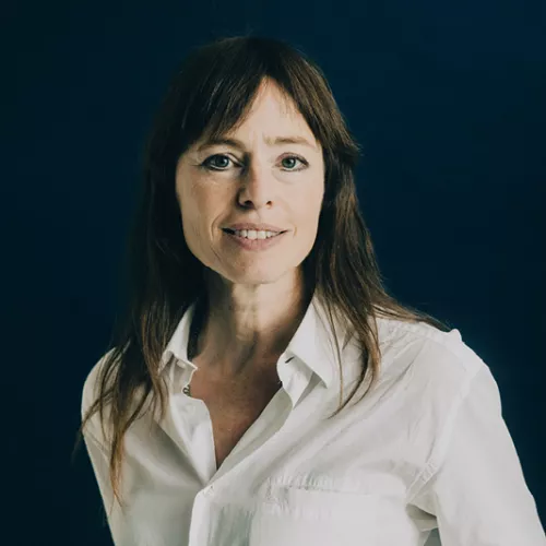 Hedwig Richter trägt lange glatte Haare und trägt eine Bluse. Sie schaut in die Kamera und lächelt. Dabei steht sie vor einem dunklen Hintergrund.